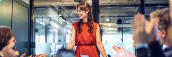 Photo of a woman standing in a meeting room being applauded by her colleagues