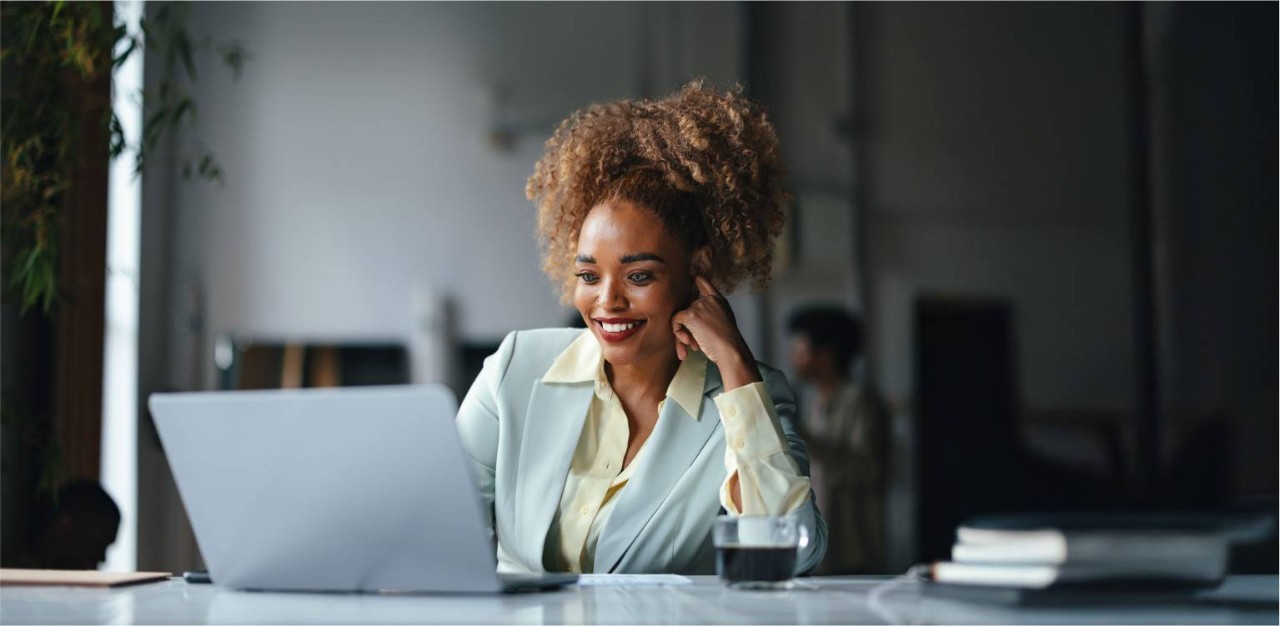 Photo of a person smiling while looking at their computer with a cup of coffee beside them 