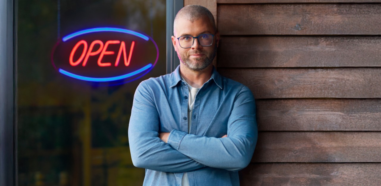 Photo of a business owner in front of their business with an open sign representing National Bank’s business transfer services