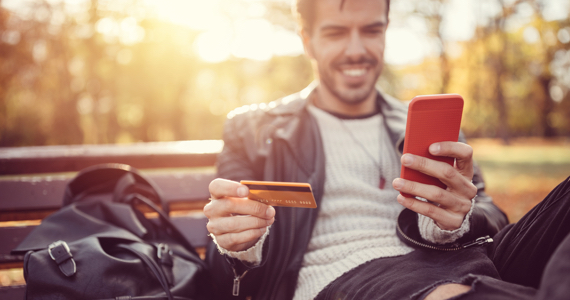 Young man sitting on a bench holds a credit card and a phone
