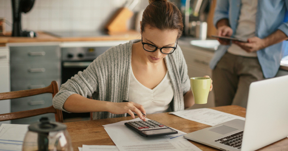 Young woman sitting at her kitchen table doing her accounts with her calculator