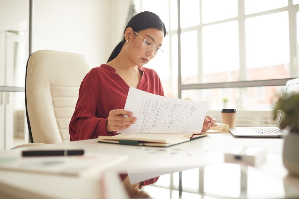 A woman siting at her desk consults papers. 