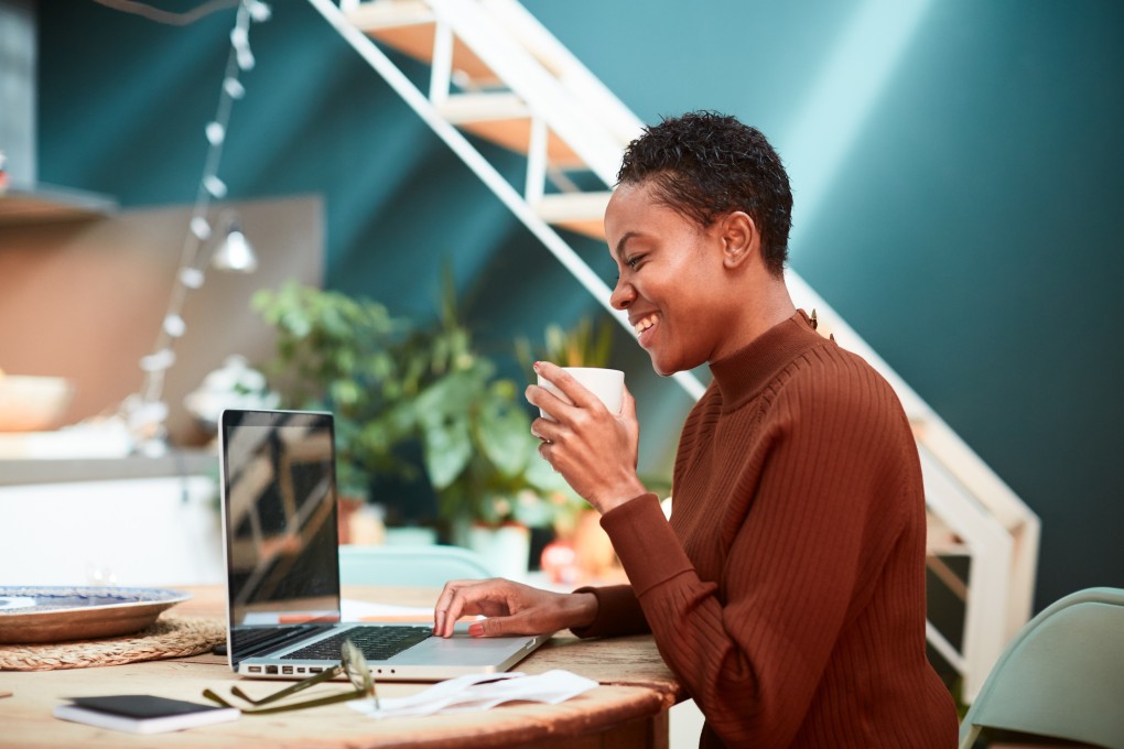 A woman working from home takes her coffee while smiling at her computer.