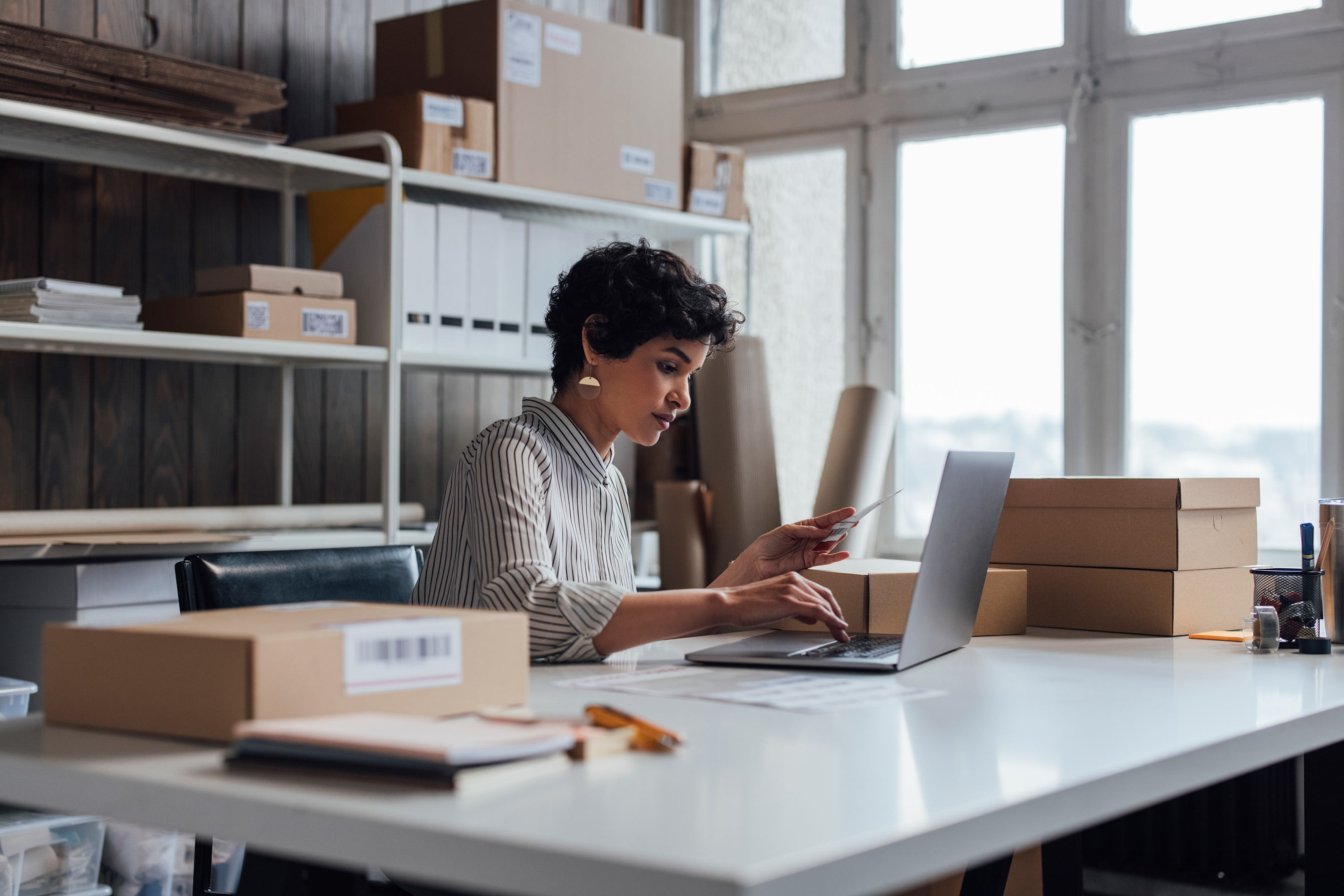 Person working on a laptop at a desk with cardboard boxes in a bright office. 