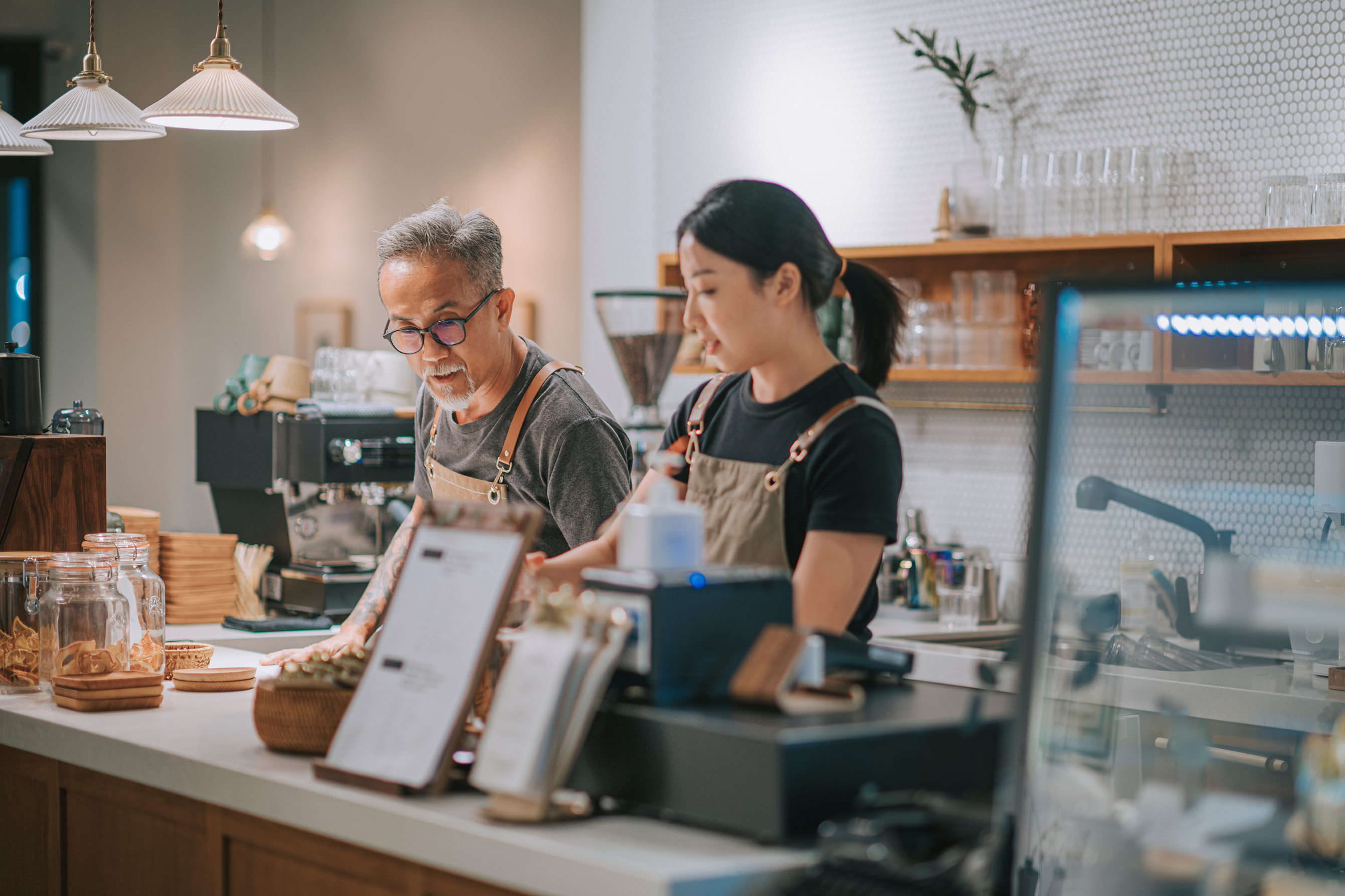 Coffee shop counter with business owner and daughter working, espresso machine and pastries in view. 