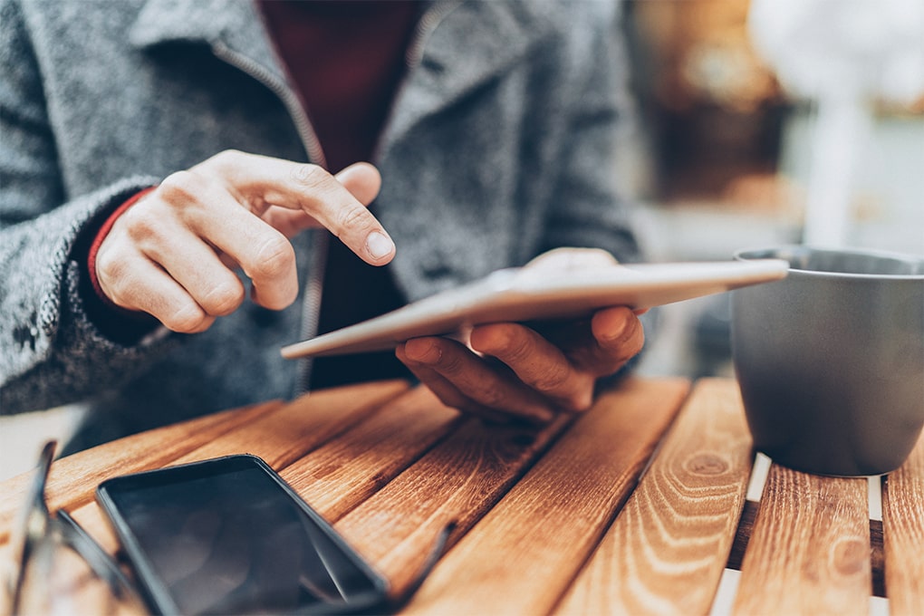 Image of a man's hands using a tablet on a table with a cup and a cell phone.   